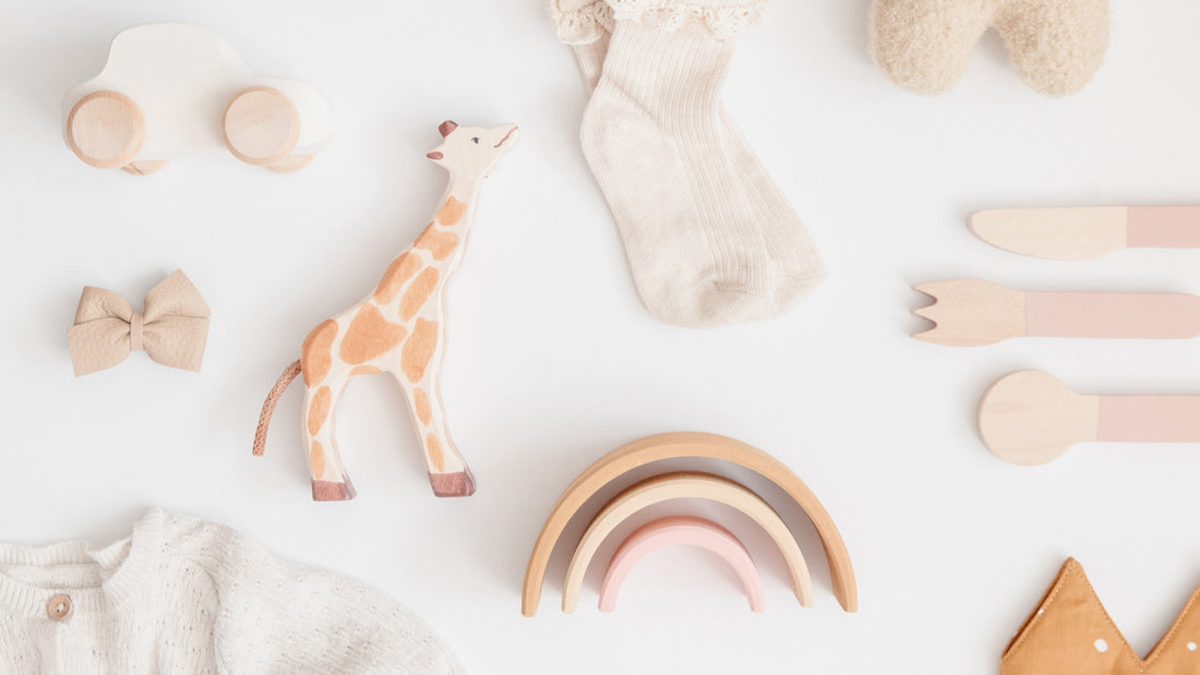 Verschiedene Geschenke zur Geburt, darunter ein Holzspielzeug in Form einer Giraffe, eine gestrickte Babydecke, Babykleidung und Holzgeschirr, liebevoll arrangiert auf einer weißen Oberfläche